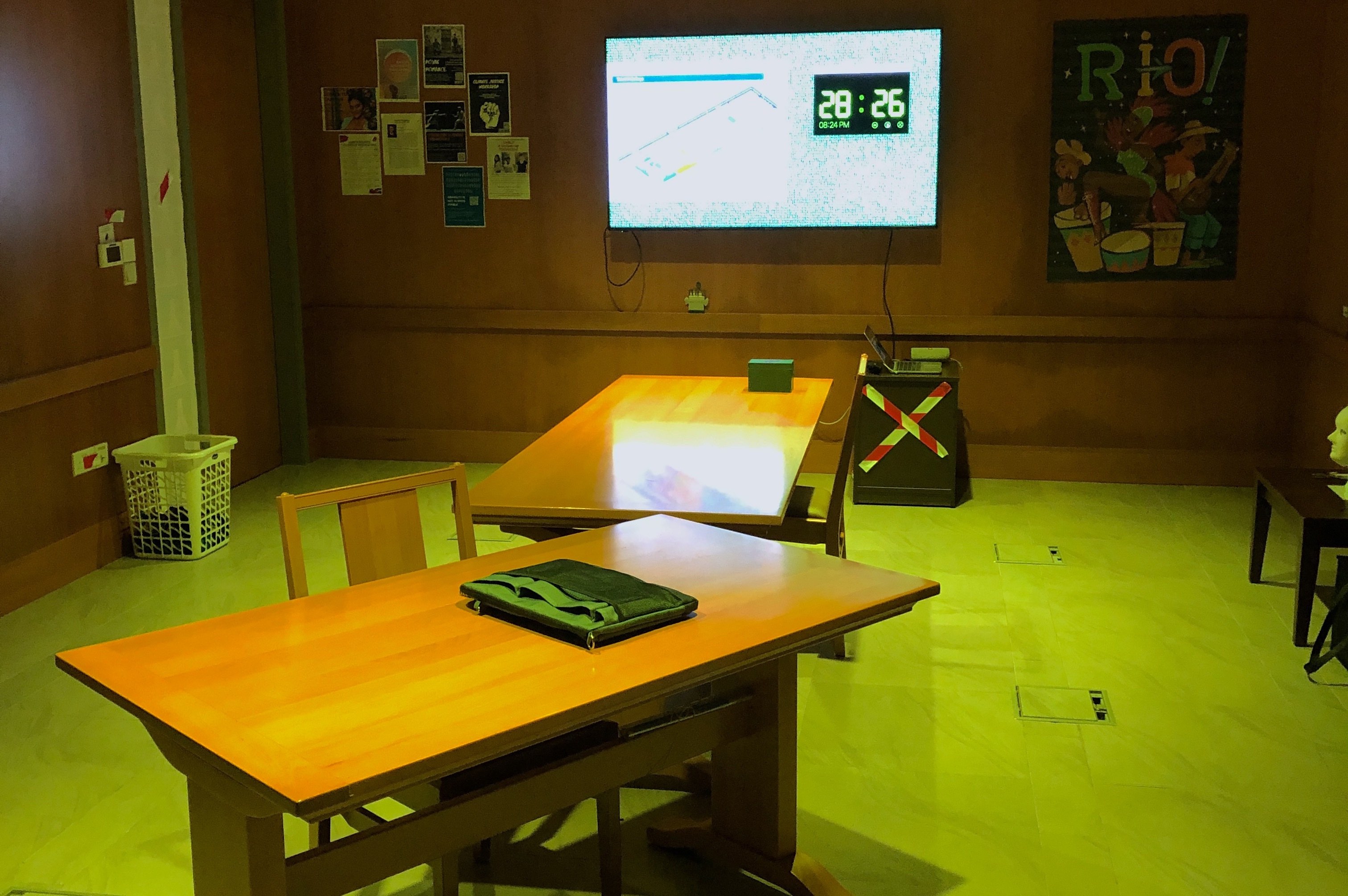 Photograph of a vacant room with green lights. Two tables with a locked pouch on them. A TV showing a timer, 28 minutes 26 seconds. There are many posters on the walls.