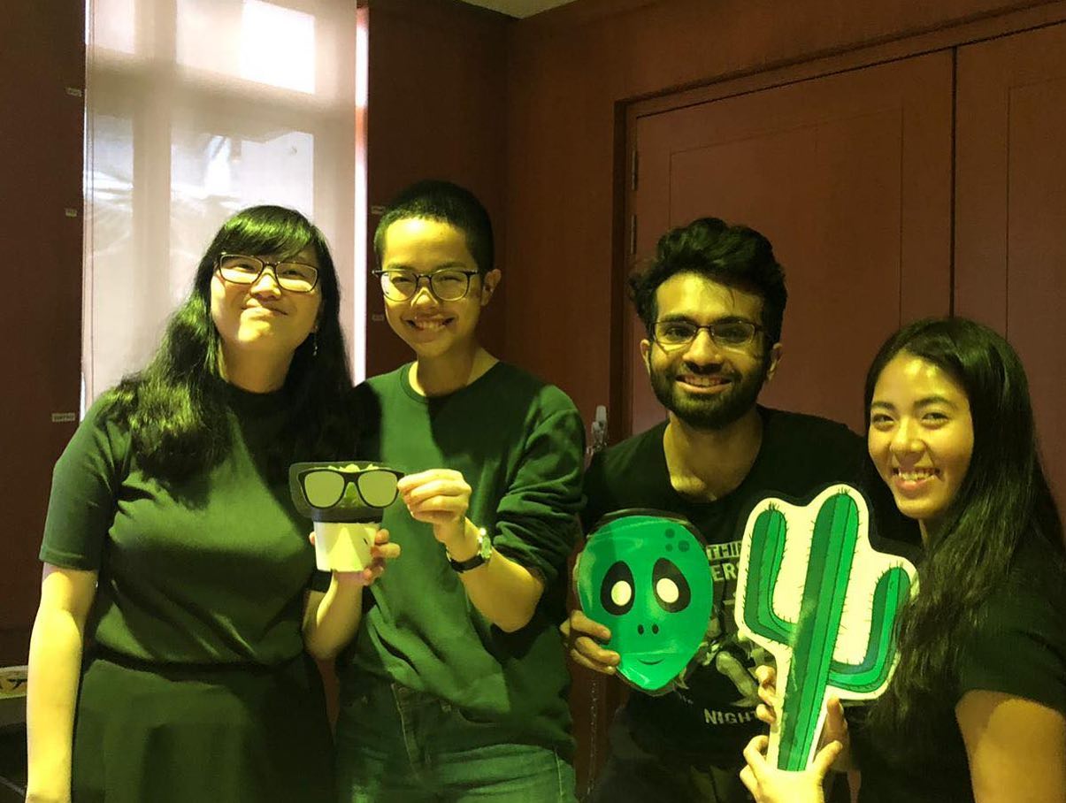 Photograph of four smiling students holding props. From left to right, a small potted plant. Paper spectacles. A printed alien mask. Cartoon drawing of a cactus.