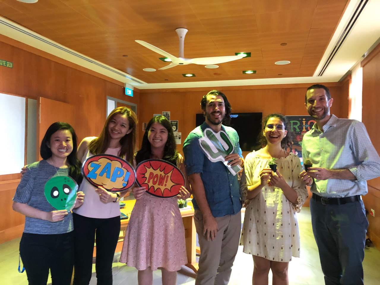 Photograph of six smiling staff members holding props. From left to right, a printed alien mask. Boards with ZAP and POW written on them. A cartoon drawing of a cactus. Two potted cactuses.